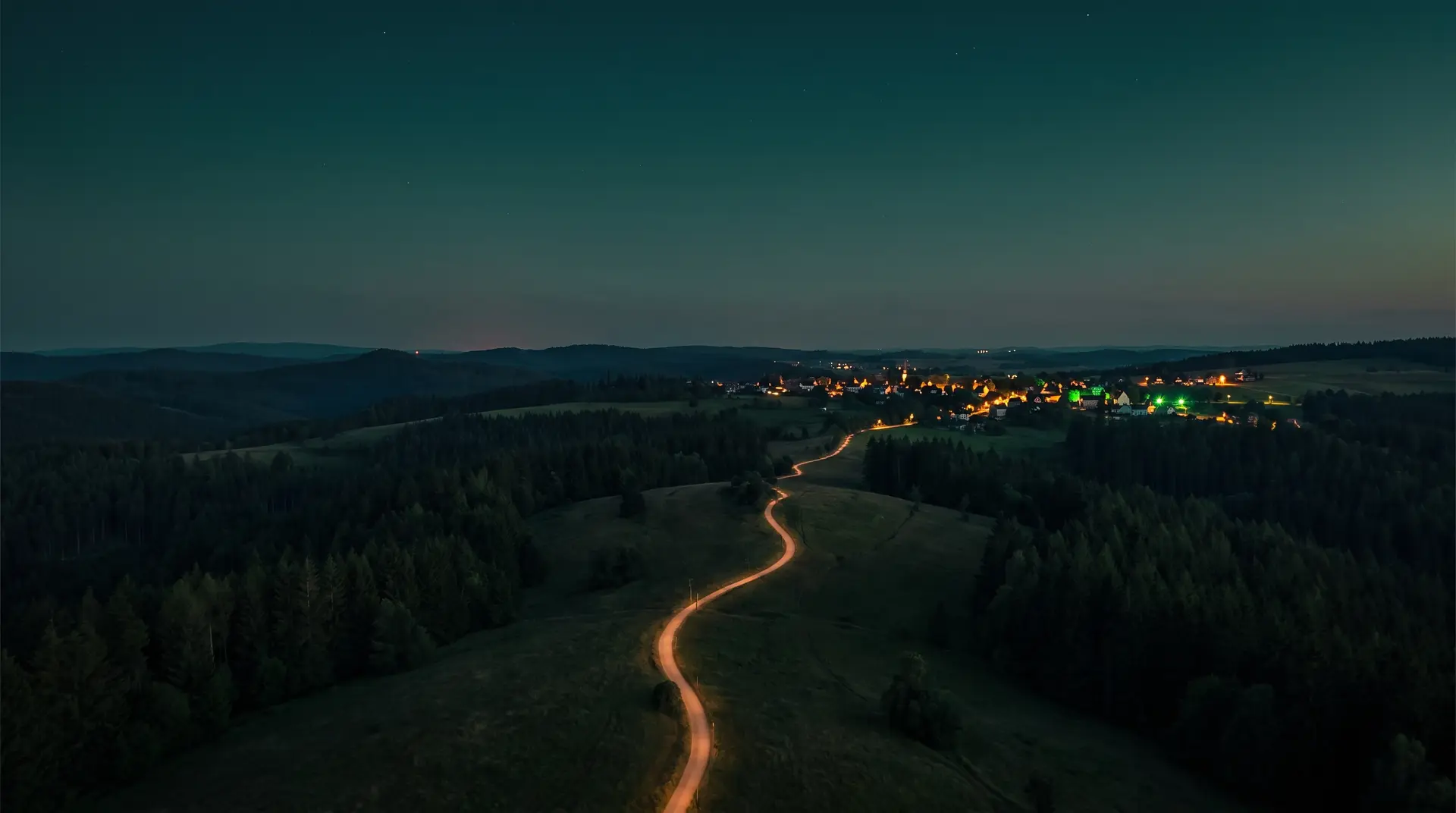 A winding footpath lit like a golden thread, leading across dark evening hills to the warm lights of a distant town